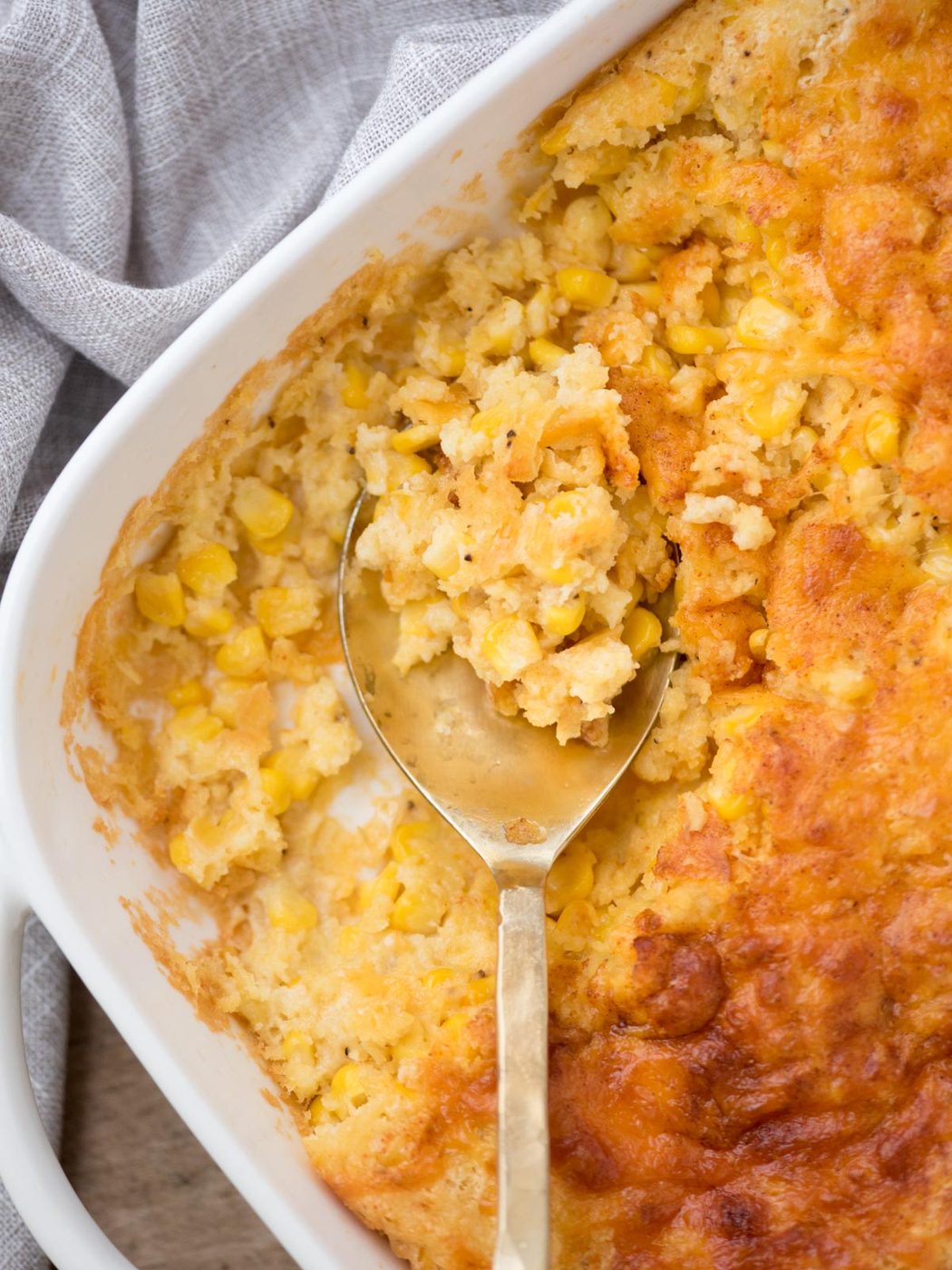A close-up of a baked corn casserole in a white dish. The golden crust is partially scooped out with a metal spoon, revealing a creamy mixture of corn and breading. A gray cloth is draped beside the dish.