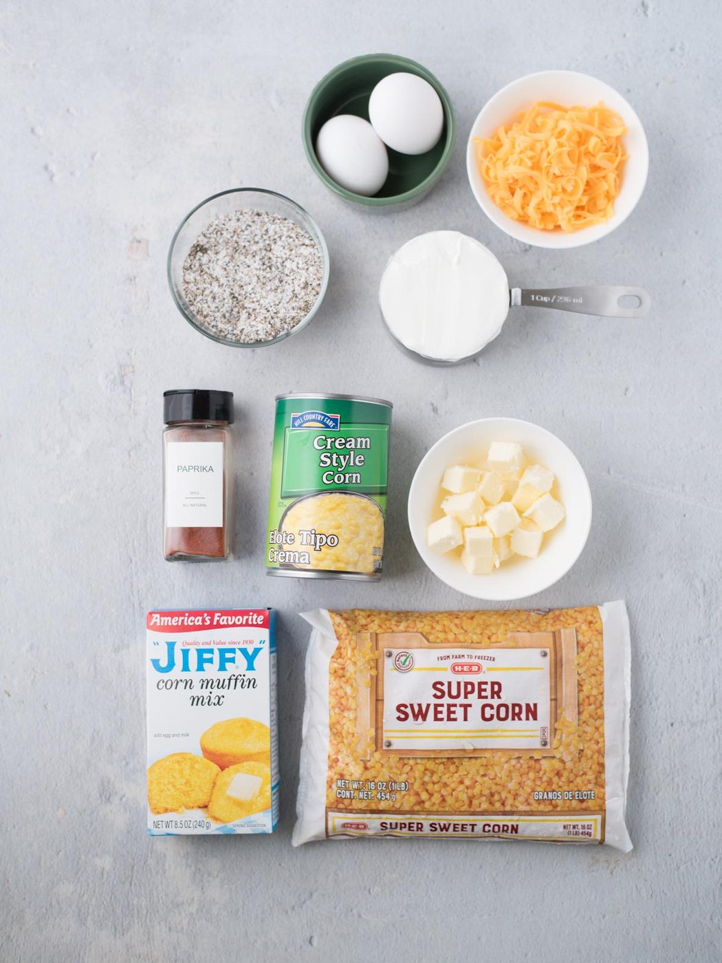 Assorted ingredients on a table: a can of creamed corn, a bag of super sweet corn, Jiffy corn muffin mix, cheddar cheese, butter cubes, two eggs, a measuring cup of sugar, a bowl of black pepper, and paprika.