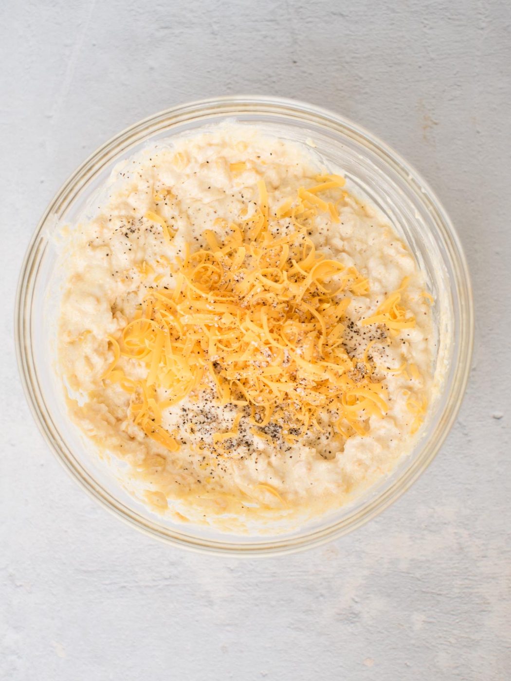 A glass bowl filled with a southern corn casserole mix, is topped with shredded cheddar cheese and black pepper, sitting on a light gray surface.