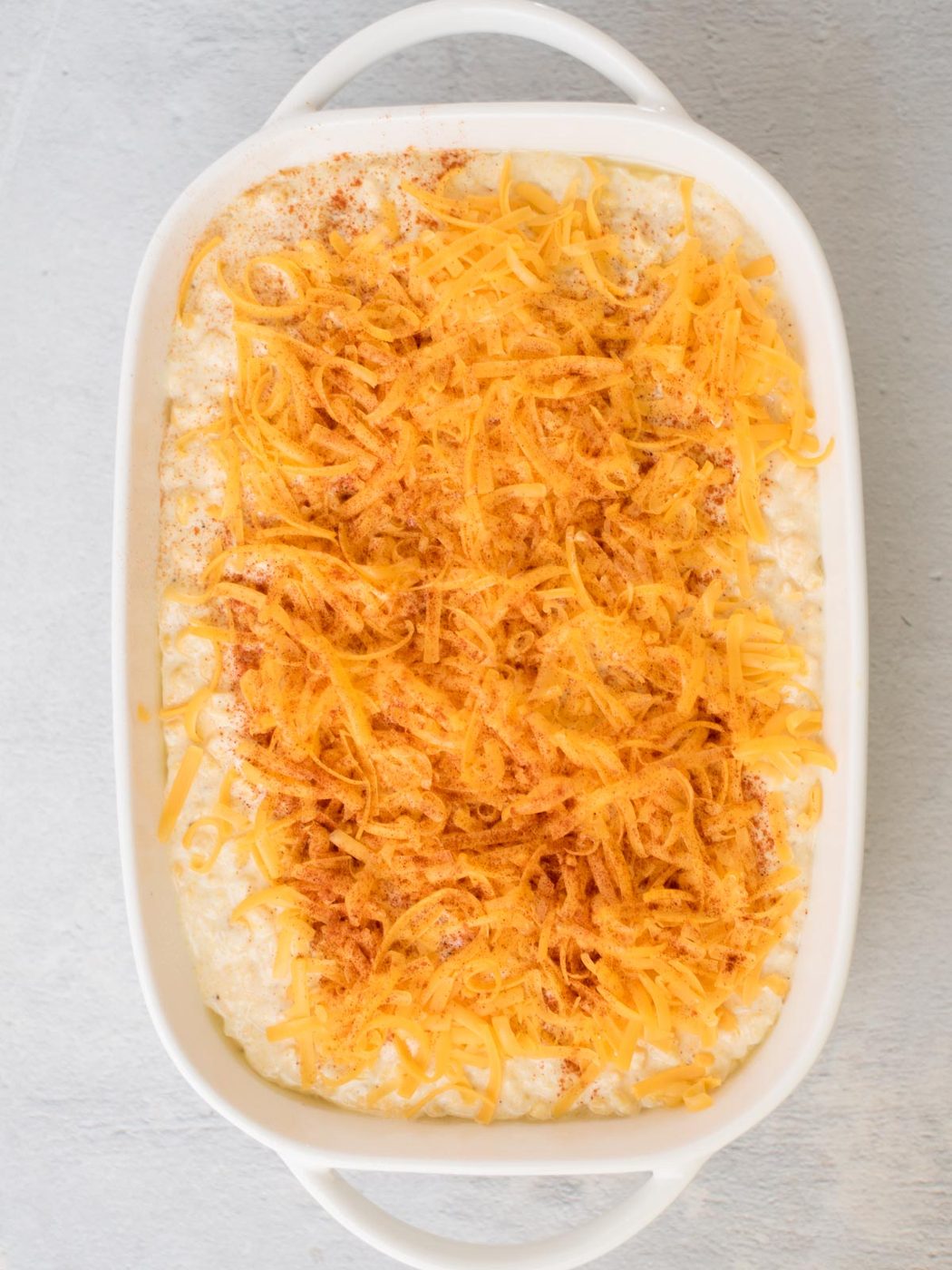 A white casserole dish filled with a creamy, old-fashioned corn casserole mixture, topped with shredded cheese and sprinkled with paprika or a similar seasoning, placed on a light gray surface.