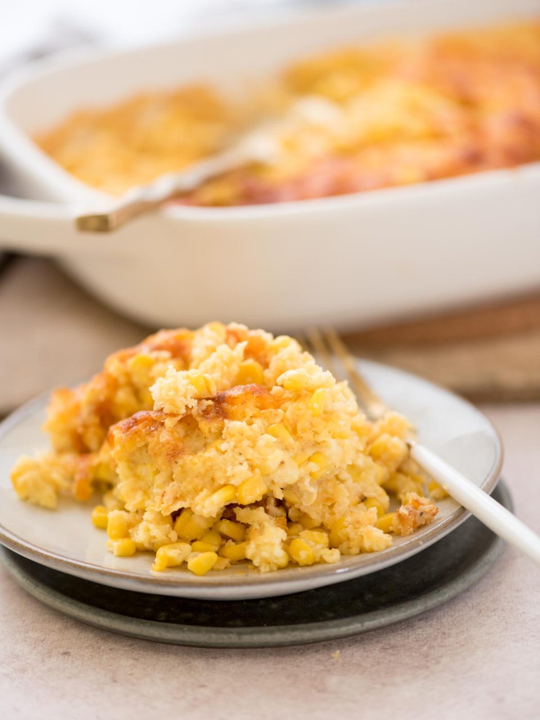 A serving of corn casserole sits on a plate with a fork, in front of a larger dish filled with the same casserole. The dish is topped with a golden, slightly crispy crust.