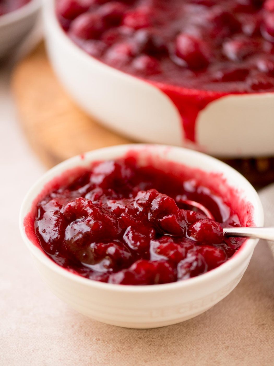 A close-up of a small white bowl filled with from-scratch cranberry sauce, showing glistening whole cranberries. A larger bowl with more sauce is in the background, slightly overflowing. A spoon is in the small bowl.