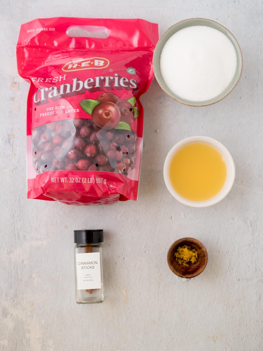 A bag of fresh cranberries is placed next to a bowl of sugar, a bowl of orange juice, a bottle of cinnamon sticks, and a small dish of lemon zest on a light countertop.