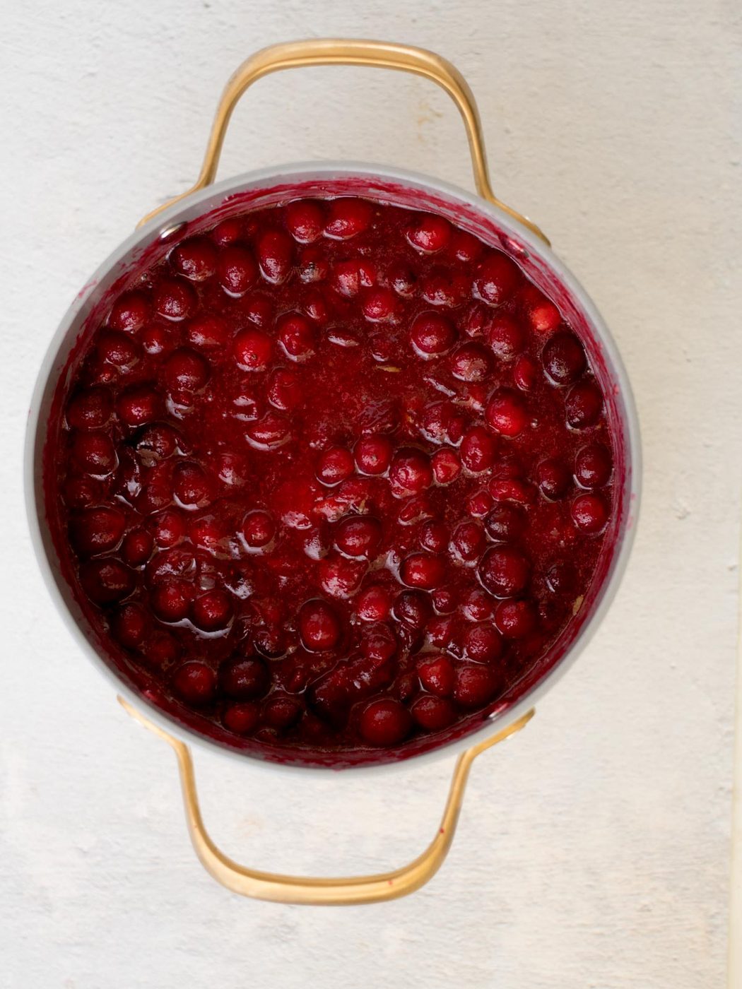 A pot filled with vibrant red cranberry sauce, featuring whole cranberries, against a light background. The pot has golden handles, adding a decorative touch to the simple presentation.