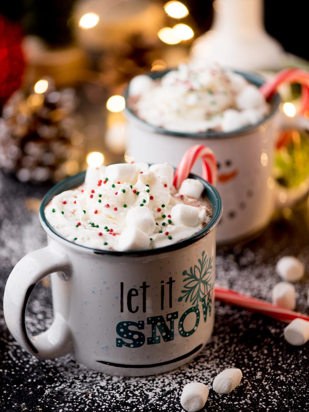 Two mugs of hot chocolate topped with whipped cream, colorful sprinkles, marshmallows, and candy canes. The front mug features the text let it snow. A festive background with fairy lights and snow-like powder completes the scene.