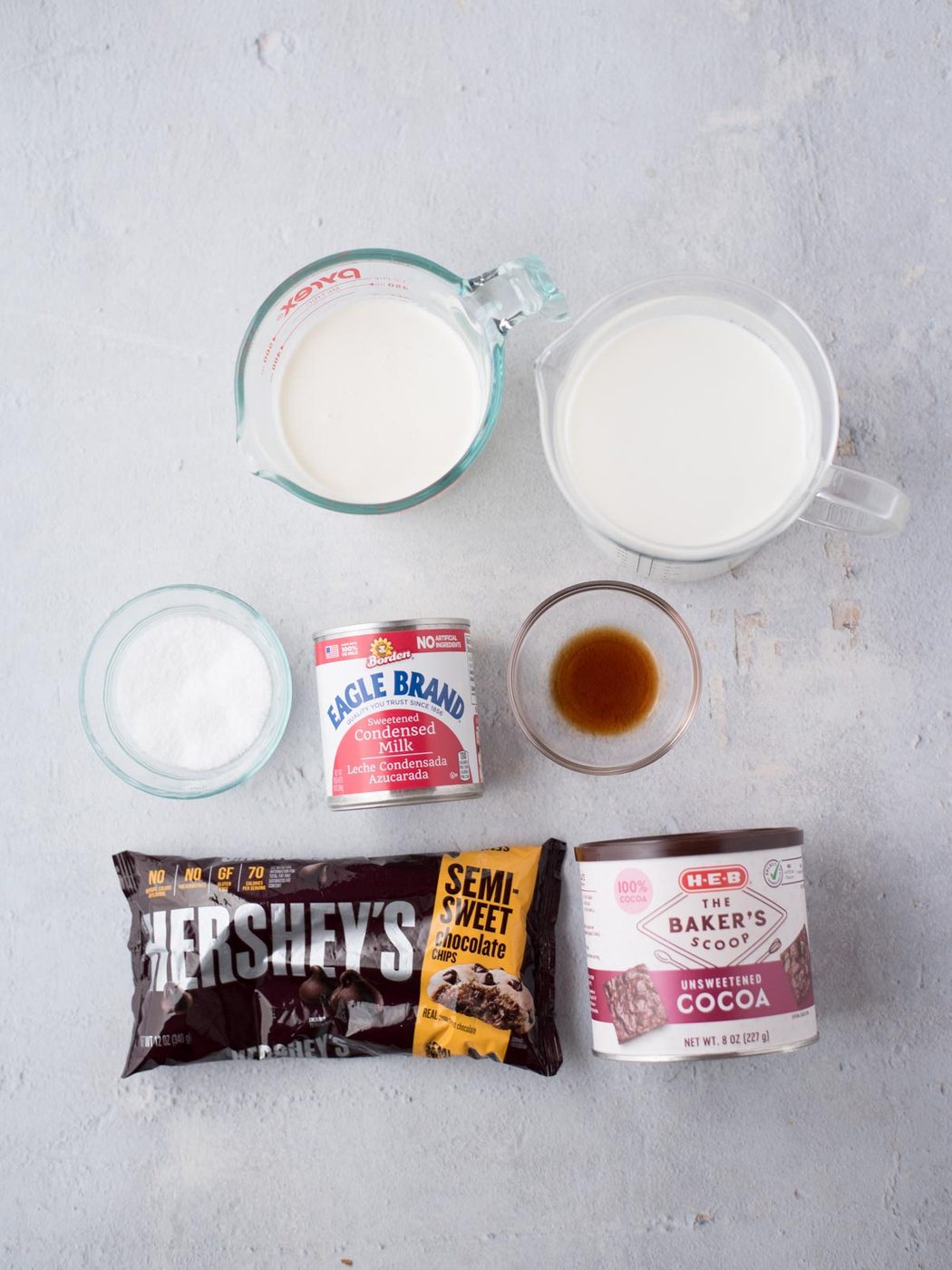 Ingredients for a dessert are laid out on a light surface: a bag of semi-sweet chocolate chips, a can of sweetened condensed milk, a small bowl of sugar, a glass of vanilla extract, a carton of heavy cream, and a box of unsweetened cocoa.