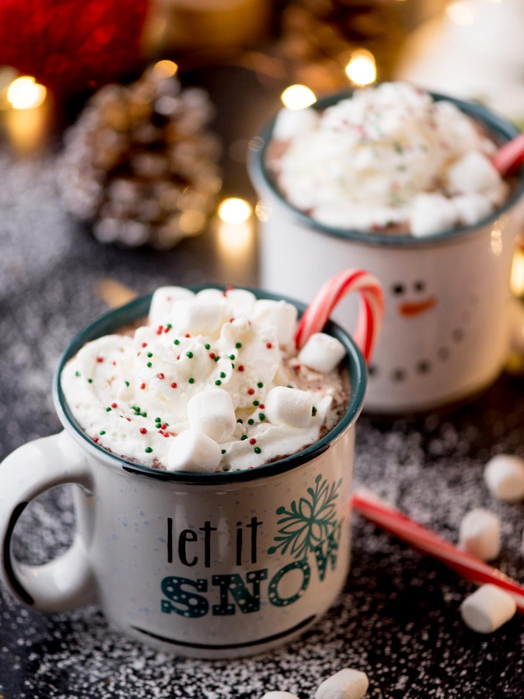 Two festive mugs of hot chocolate topped with whipped cream, sprinkles, and marshmallows. One mug has a candy cane and Let it Snow printed on it. The scene is sprinkled with powdered sugar, and there are twinkling lights and a pinecone in the background.