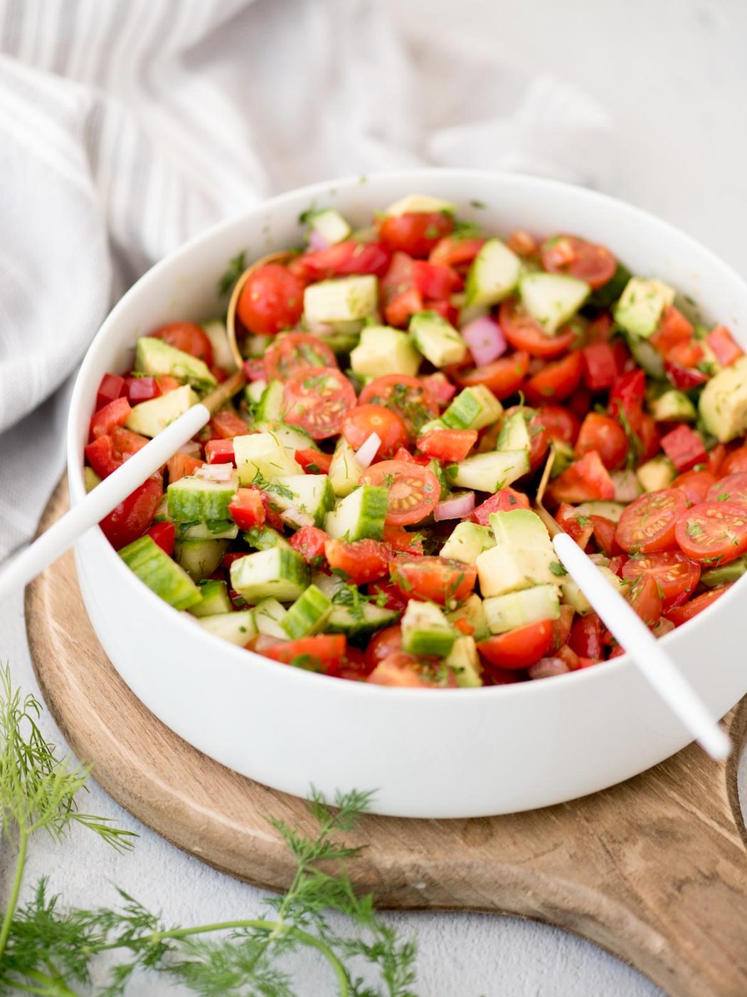 White salad bowl with cucumber tomato salad in it and salad tongs resting inside sitting on a wooden cutting board.