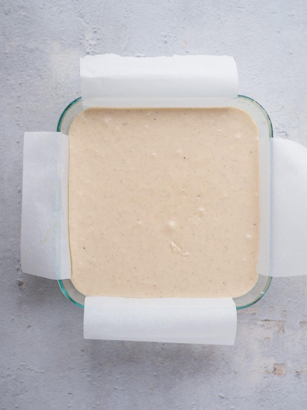 Overhead view of a square glass baking dish lined with parchment paper, filled with light beige eggnog fudge batter, set on a textured gray surface.