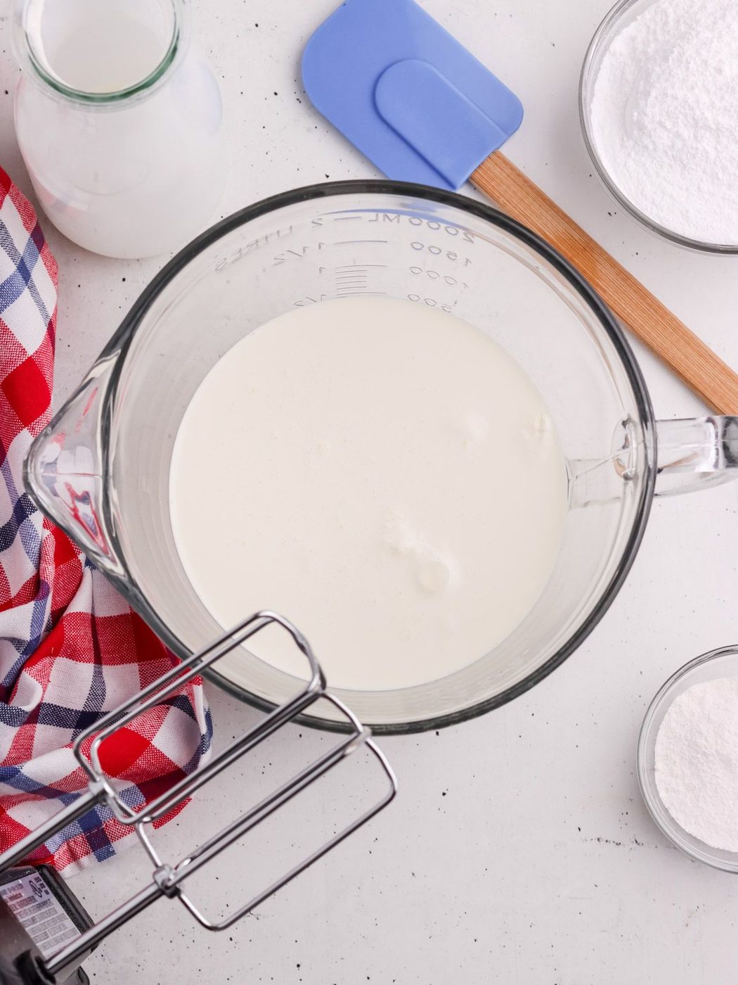 A glass mixing bowl filled with milk or cream sits on a white surface, surrounded by a checkered cloth, a bottle of milk, a blue spatula, a bowl of sugar, and a hand mixer.