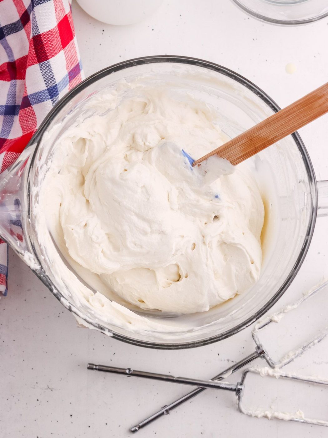 A glass mixing bowl filled with fluffy whipped cream, a wooden spatula resting on the side, and metal beaters lying nearby. A red, white, and blue checkered cloth is partially visible to the left.
