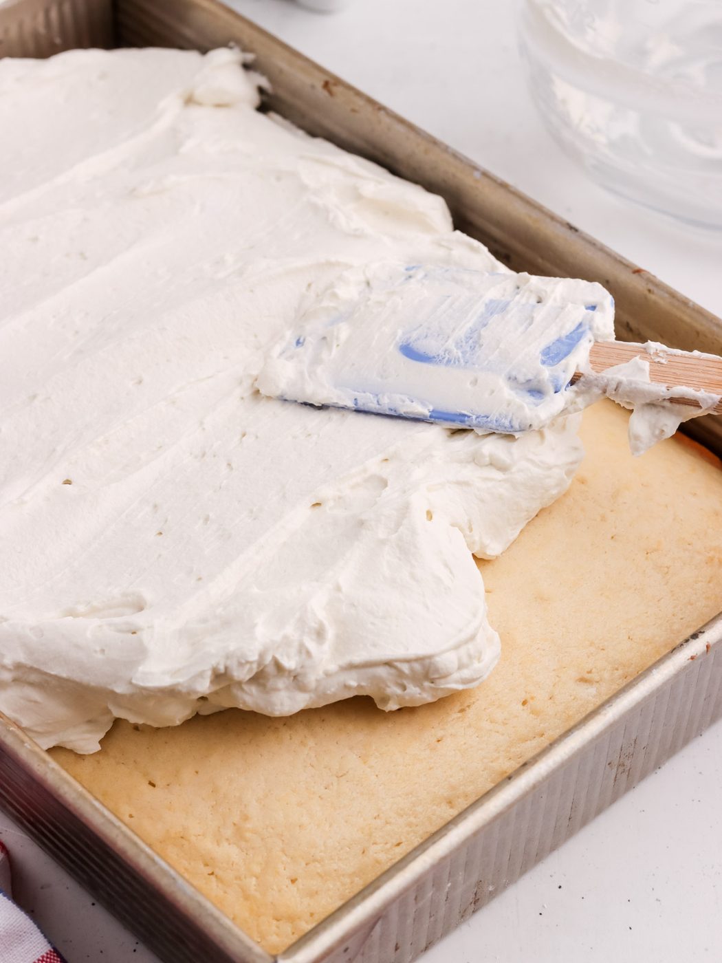 A metal baking pan holds an American Flag Cake, as a thick layer of white icing is smoothed over the rectangular cake with a blue and white spatula.
