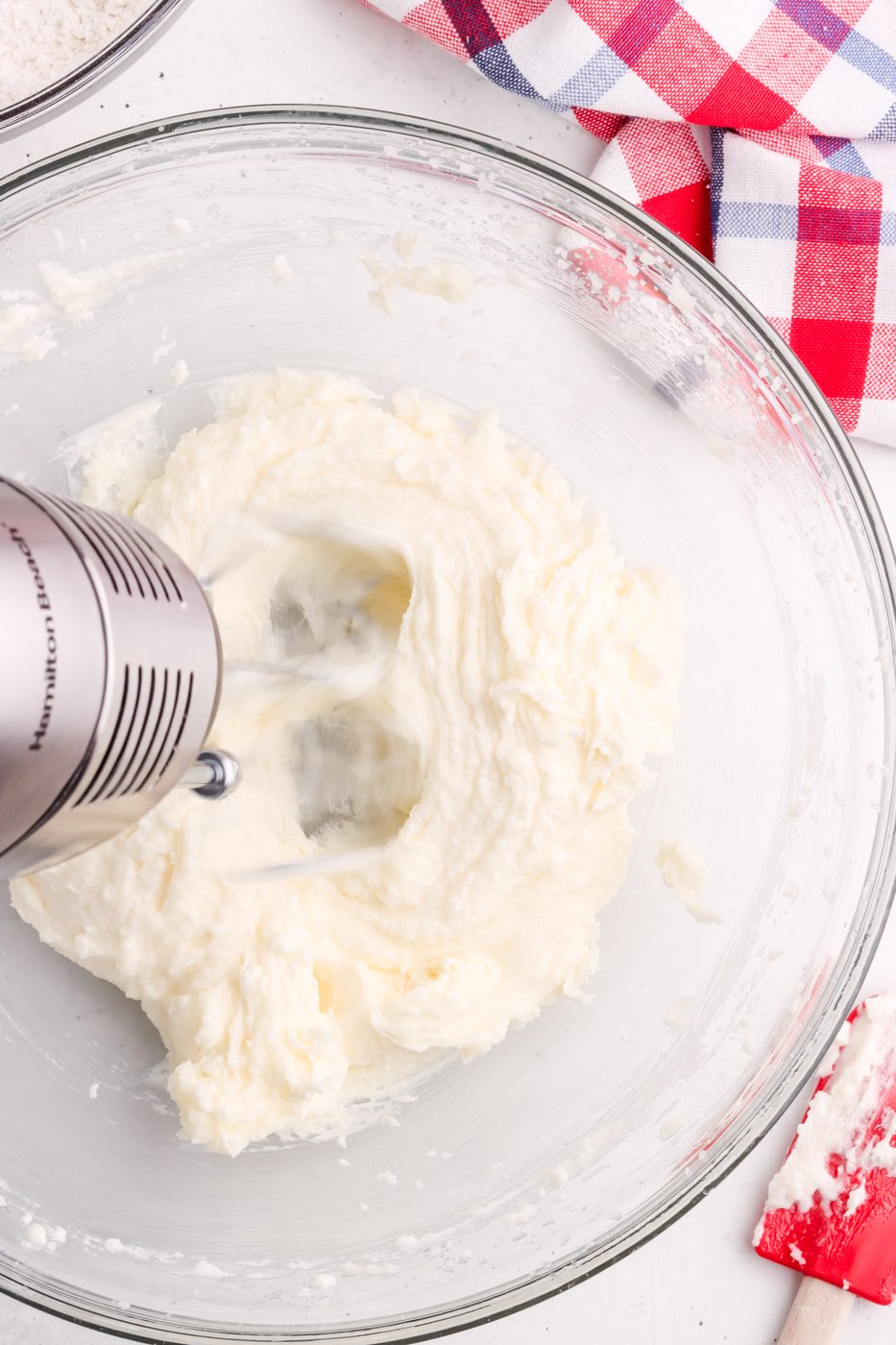 A hand mixer blends a creamy white mixture in a glass bowl, surrounded by a red and blue checkered cloth and a red spatula on a white surface.