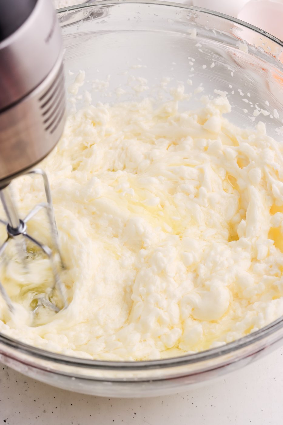 A hand mixer blending a creamy, pale mixture in a glass bowl, likely butter and sugar being whipped together on a white countertop.
