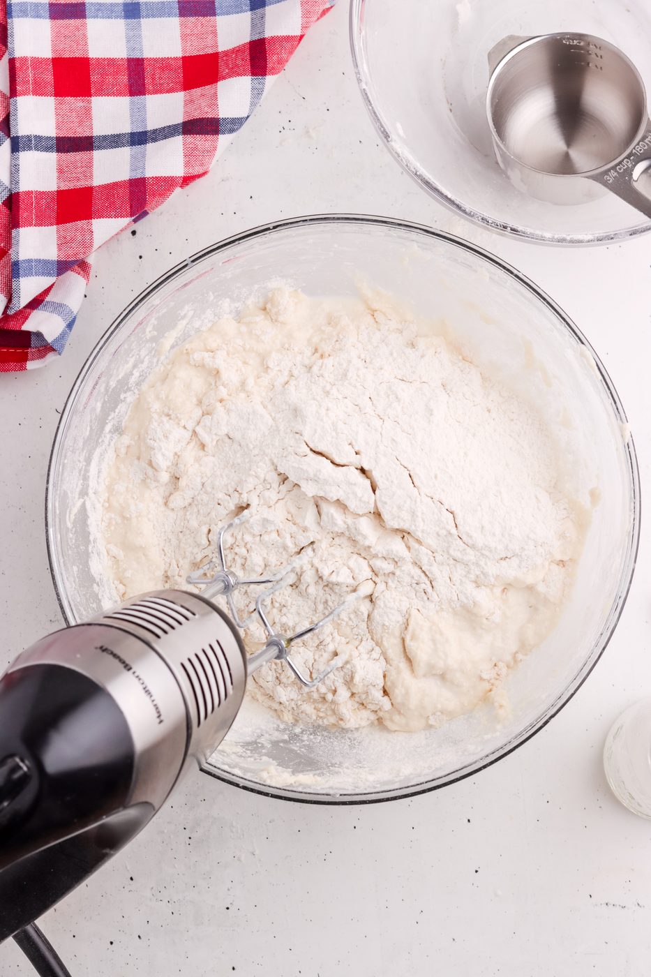 A glass bowl with flour and dough being mixed by an electric hand mixer sits on a white surface near a red, white, and blue checkered cloth and a metal measuring cup.