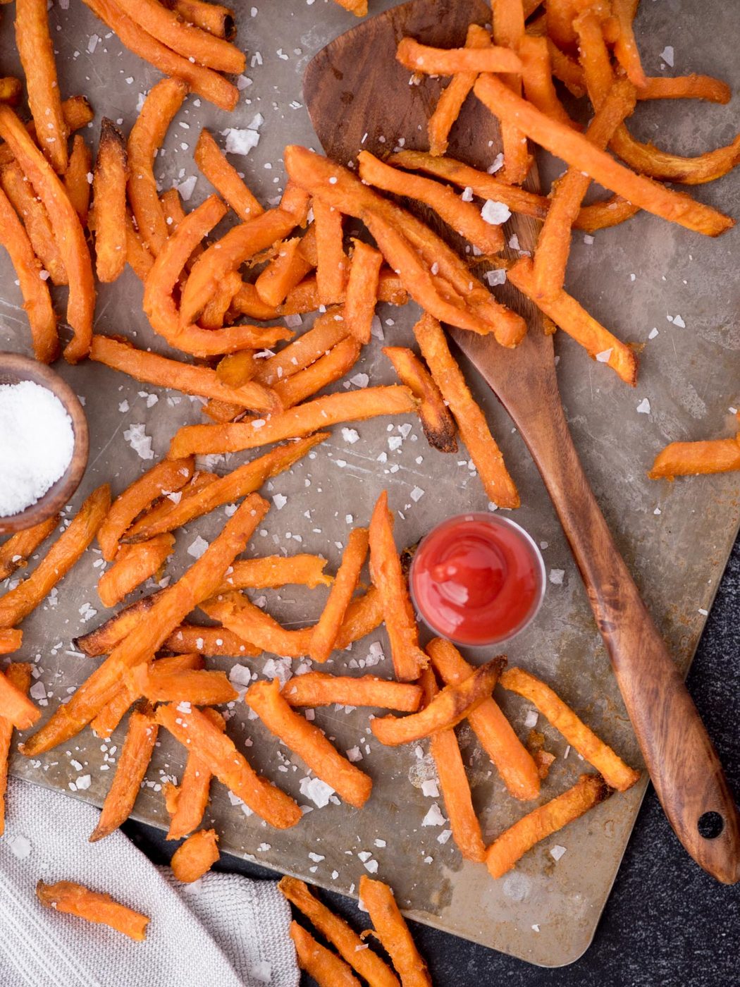 Wooden spoon laying amongst sweet potato french fries on a baking sheet and sea salt sprinkled over the top.