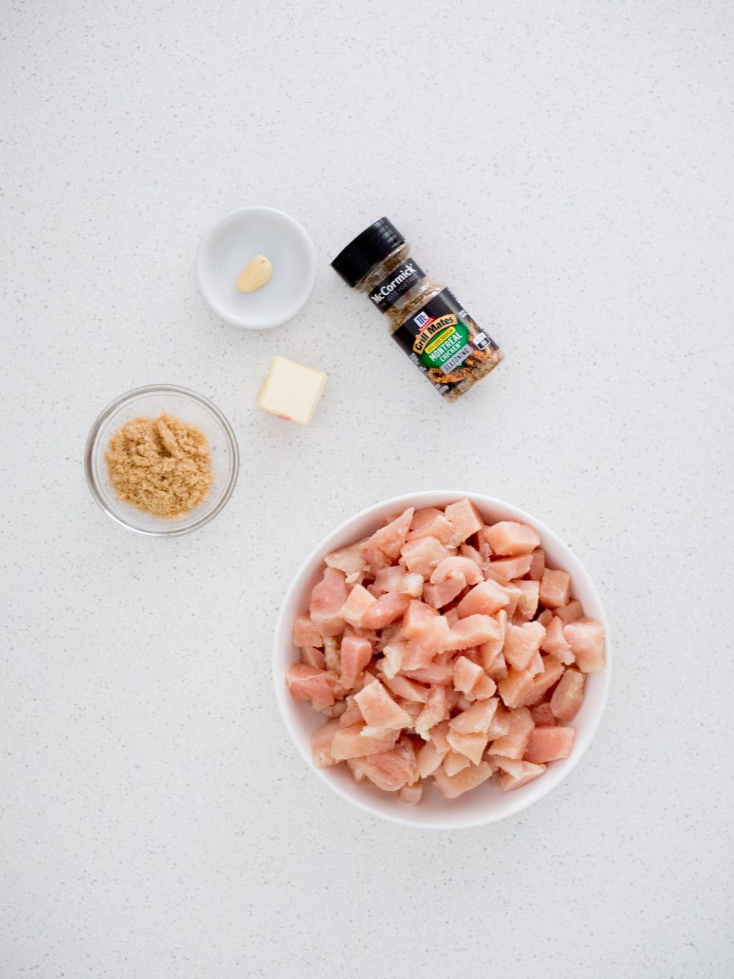 A bowl of raw, diced chicken, a small dish of brown sugar, a pat of butter, a peeled garlic clove, and a bottle of seasoning are arranged on a white countertop.
