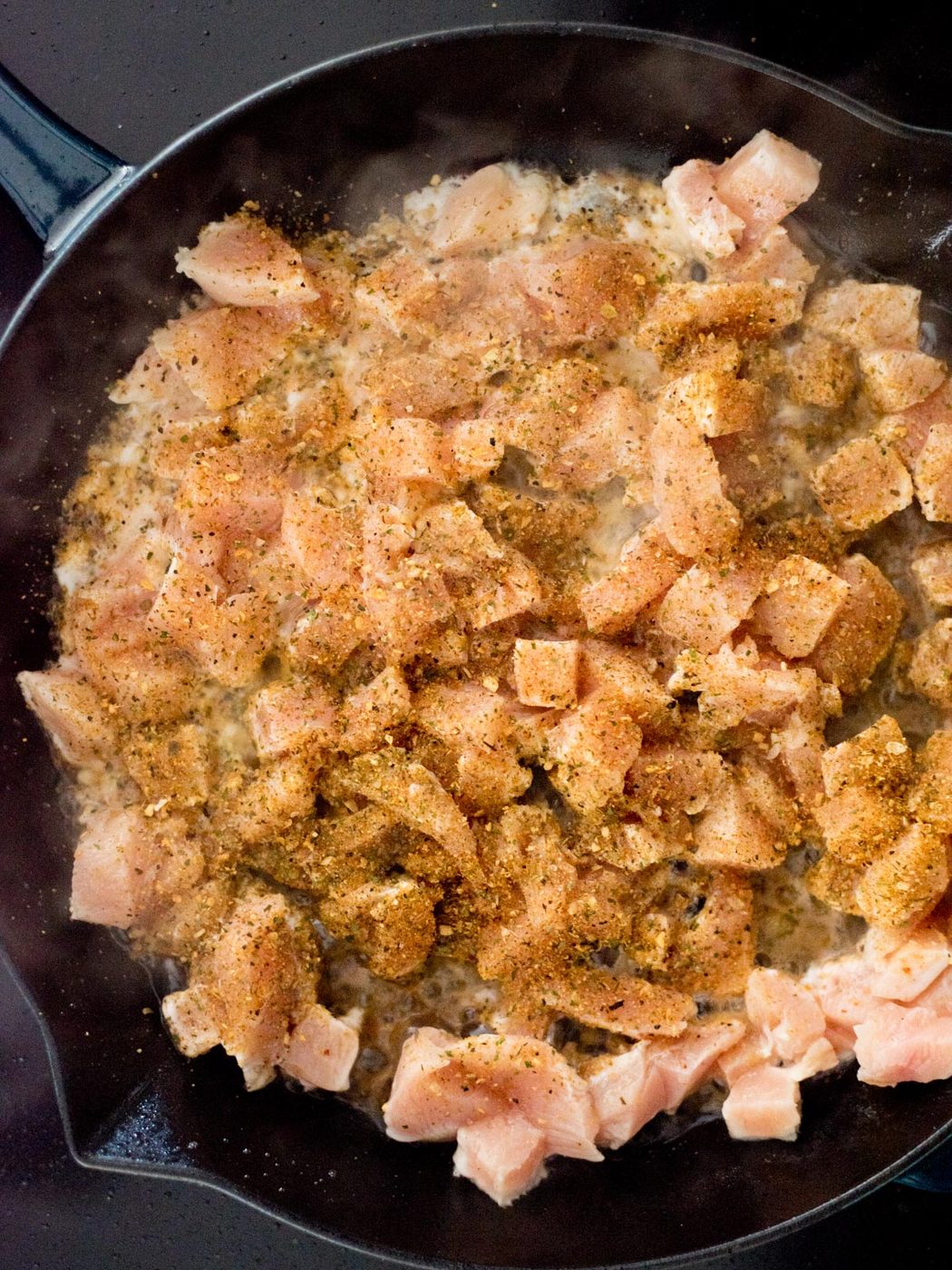 Diced raw chicken pieces in a black skillet, sprinkled with various seasonings, beginning to cook.