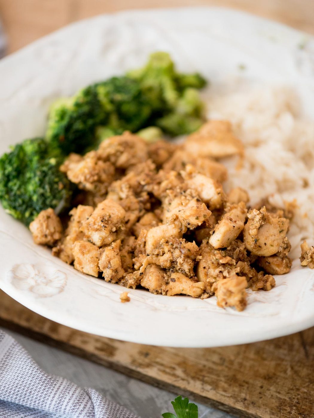 A white plate with cooked chicken pieces, steamed broccoli, and white rice, served on a wooden surface.
