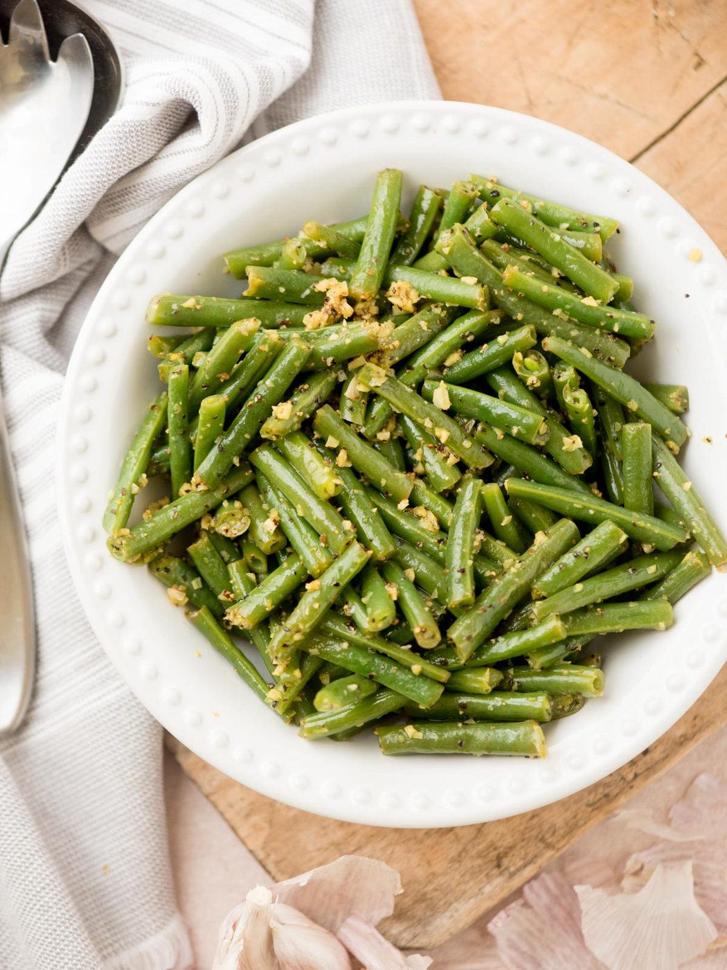 A white bowl filled with cooked green beans tossed with minced garlic and seasonings, placed on a wooden surface next to a striped towel, a metal serving spoon, and garlic cloves.