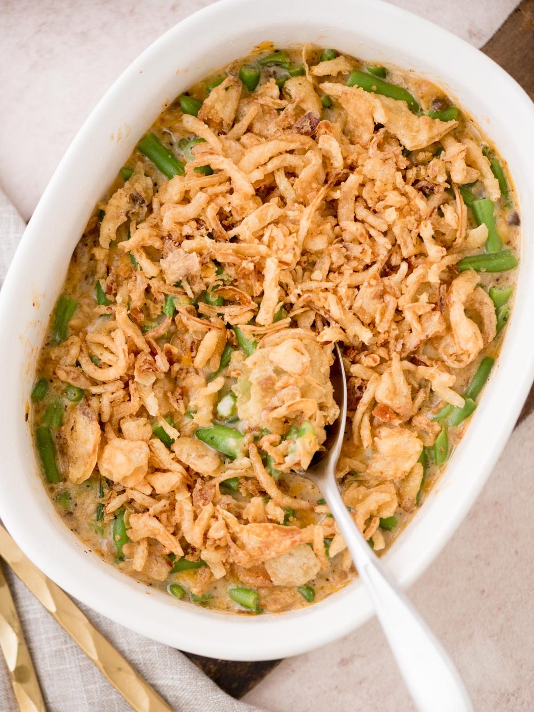 A casserole dish filled with green bean casserole topped with crispy fried onions. A spoon is placed in the dish, indicating its ready to serve. The baked dish is in a white, oval-shaped dish on a light-colored surface.