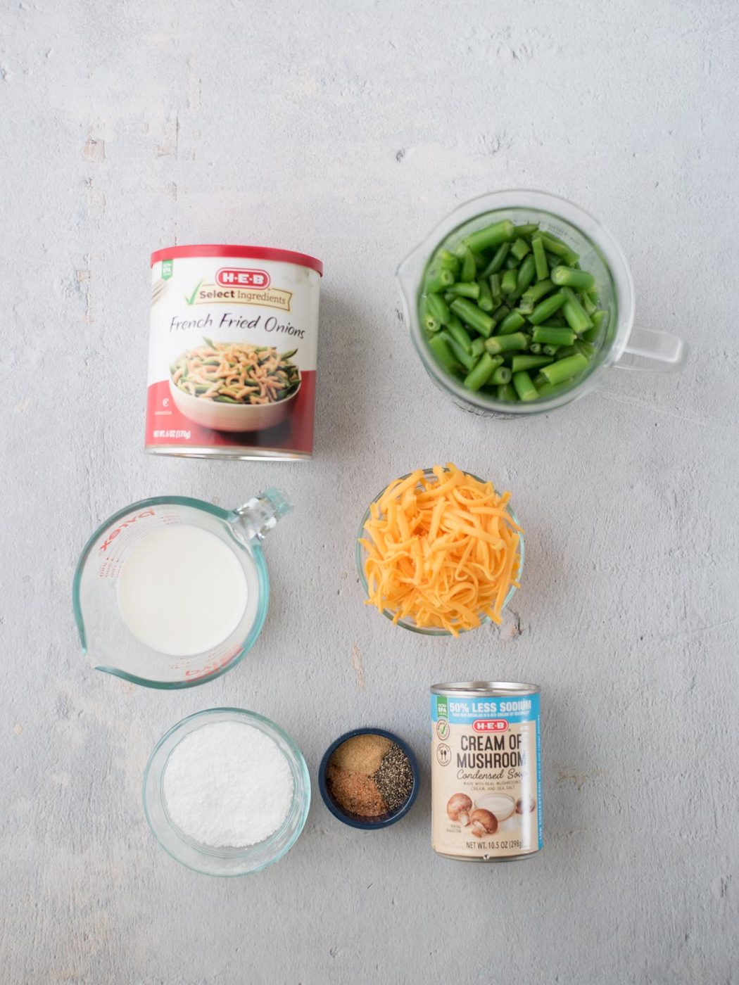Ingredients for a casserole are arranged on a surface: a can of French fried onions, a measuring cup with green beans, shredded cheese, a can of cream of mushroom soup, a measuring cup with milk, and small bowls of salt and seasoning.
