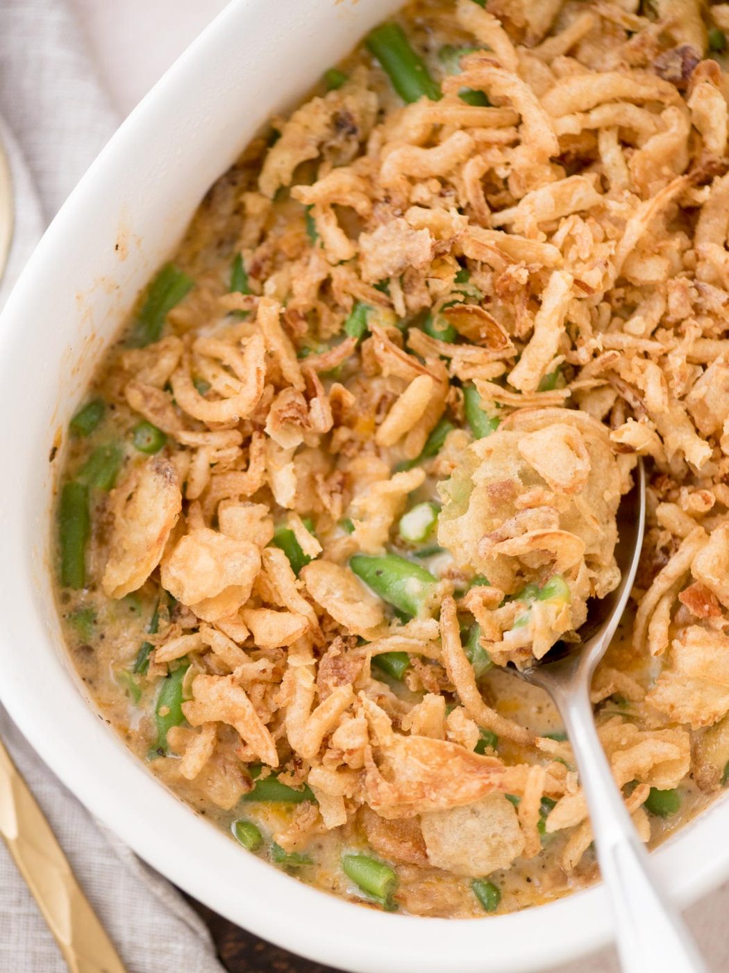 A close-up of a dish in a white casserole filled with creamy green bean casserole topped with crispy fried onions. A spoon is scooping some of the casserole, revealing the green beans and sauce beneath.