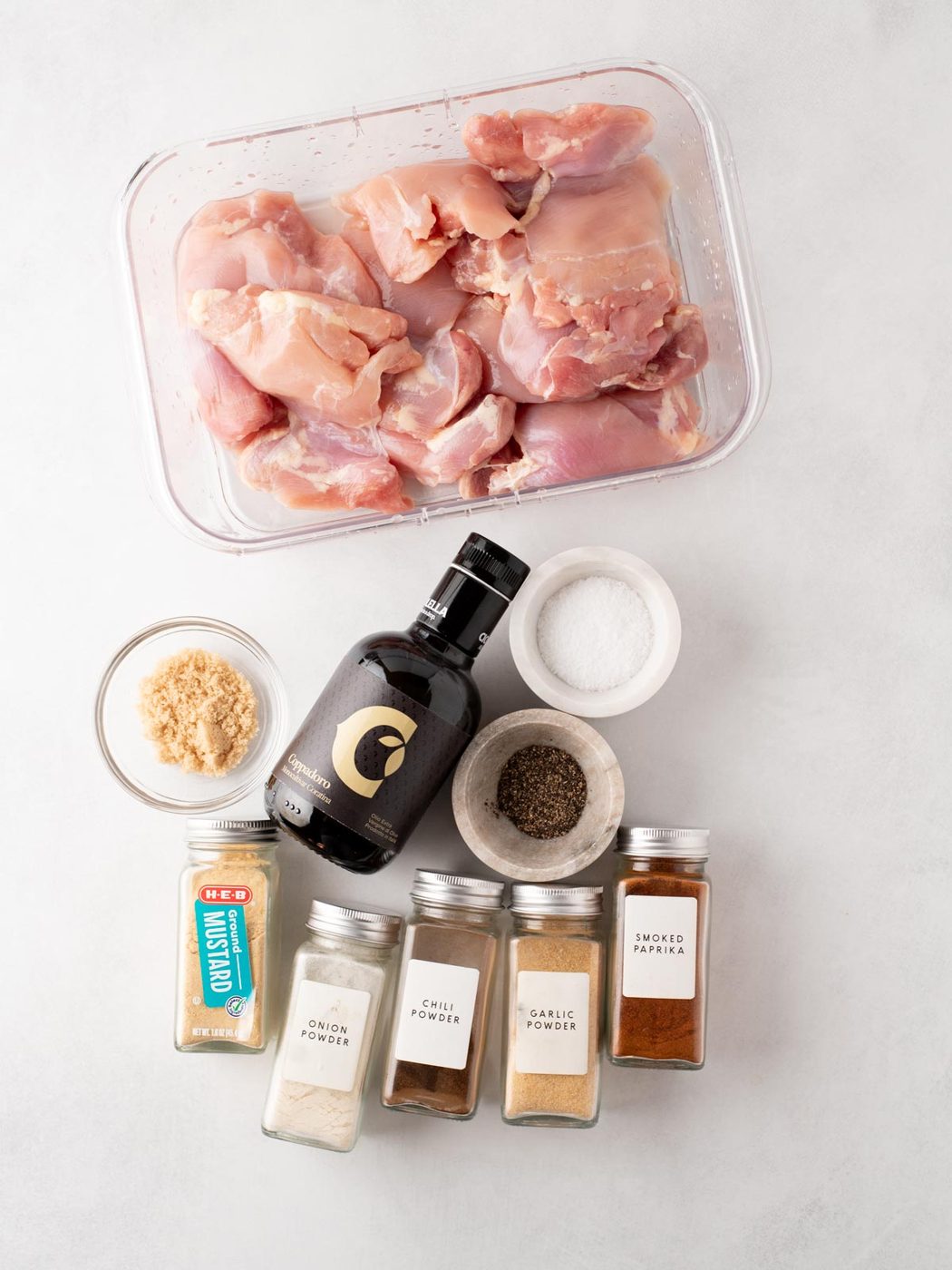 Raw chicken thighs in a glass container, surrounded by bowls and jars of ingredients including olive oil, salt, pepper, brown sugar, and various spices like mustard, onion powder, chili powder, garlic powder, and smoked paprika.