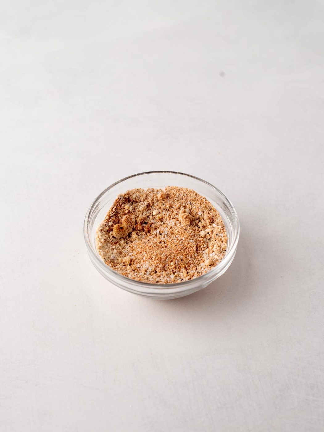 A small glass bowl filled with a light brown, finely ground seasoning mix sits on a plain, light-colored surface.