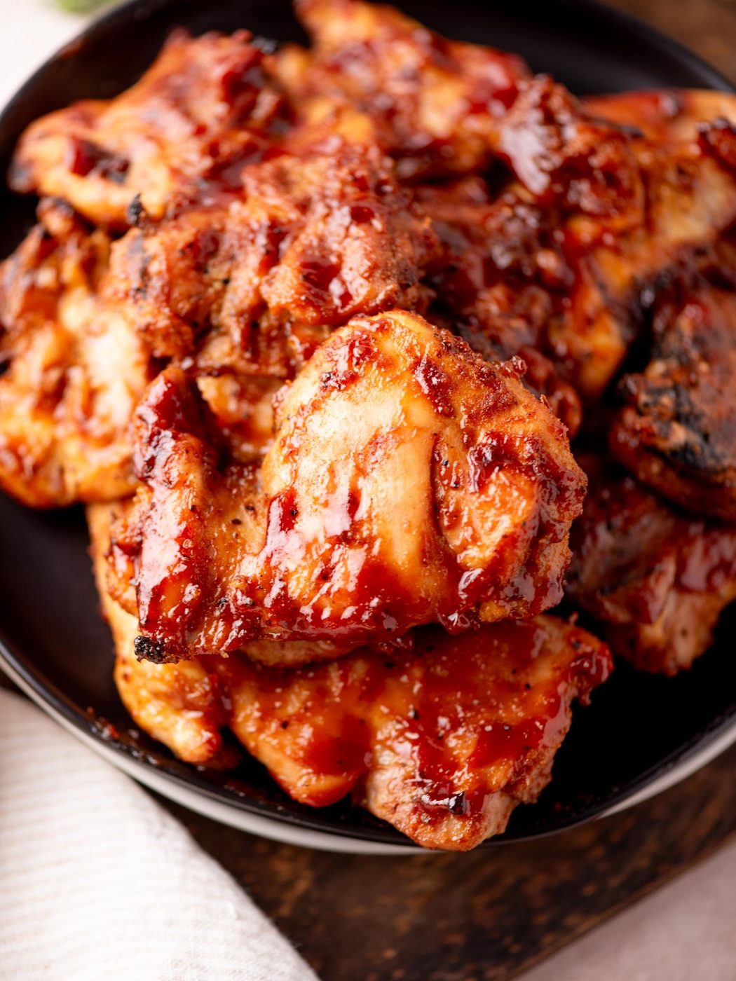 Close-up of several pieces of grilled chicken coated in a glossy, reddish barbecue sauce, stacked on a dark plate with a white cloth partially visible underneath.
