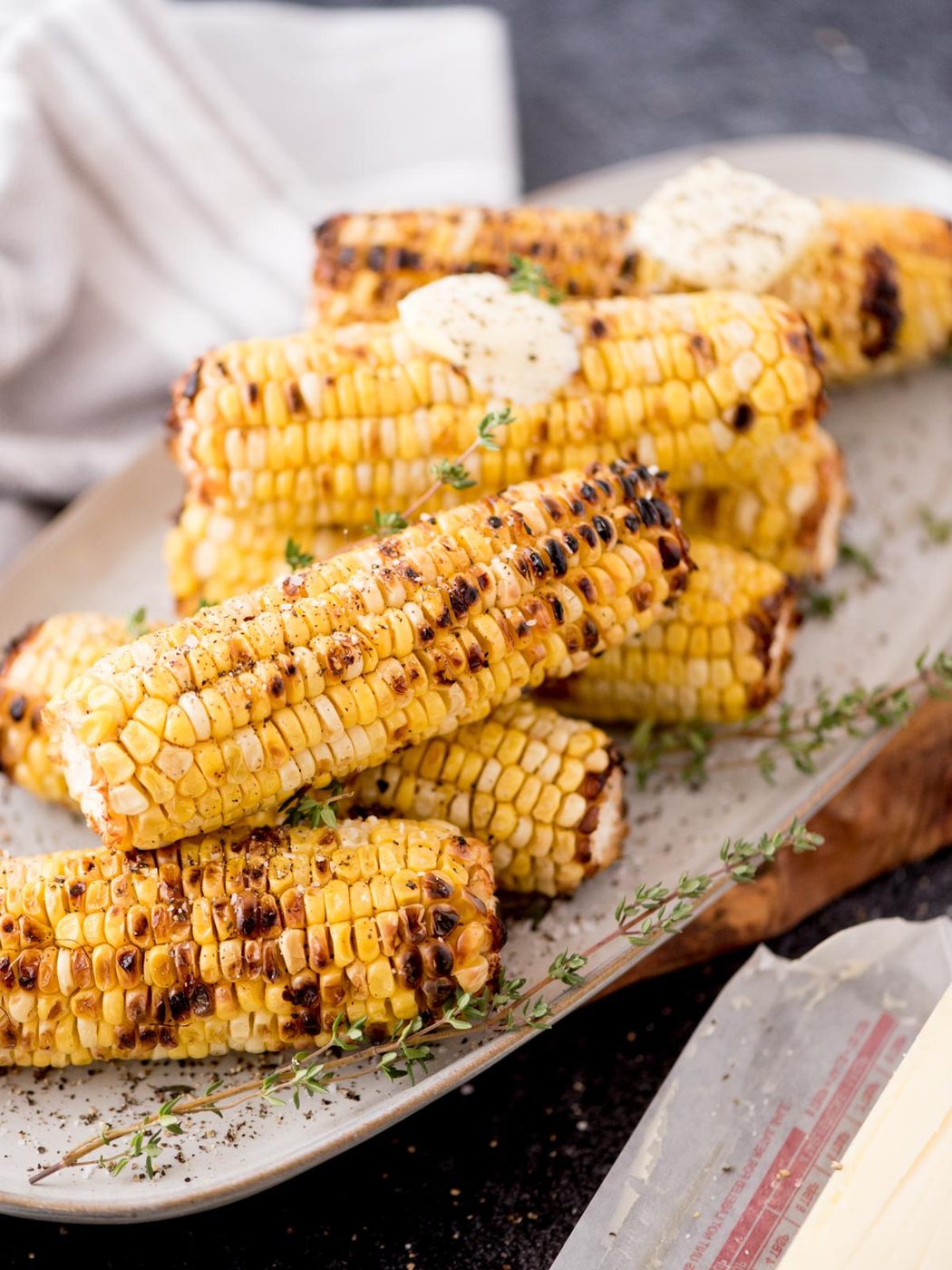 Grilled corn on the cob stacked on a plate, topped with pats of butter and garnished with fresh thyme. A white cloth napkin is in the background.