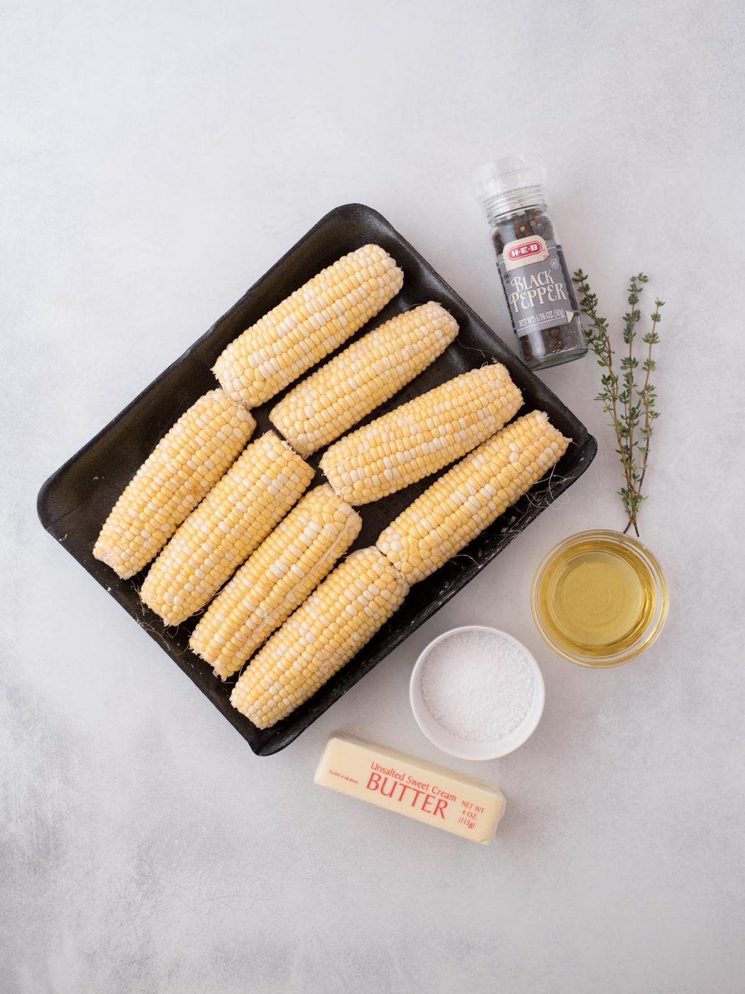 Nine ears of corn on a black tray, with a stick of butter, a bowl of salt, a small bowl of oil, a sprig of thyme, and a bottle of black pepper arranged on a white surface.