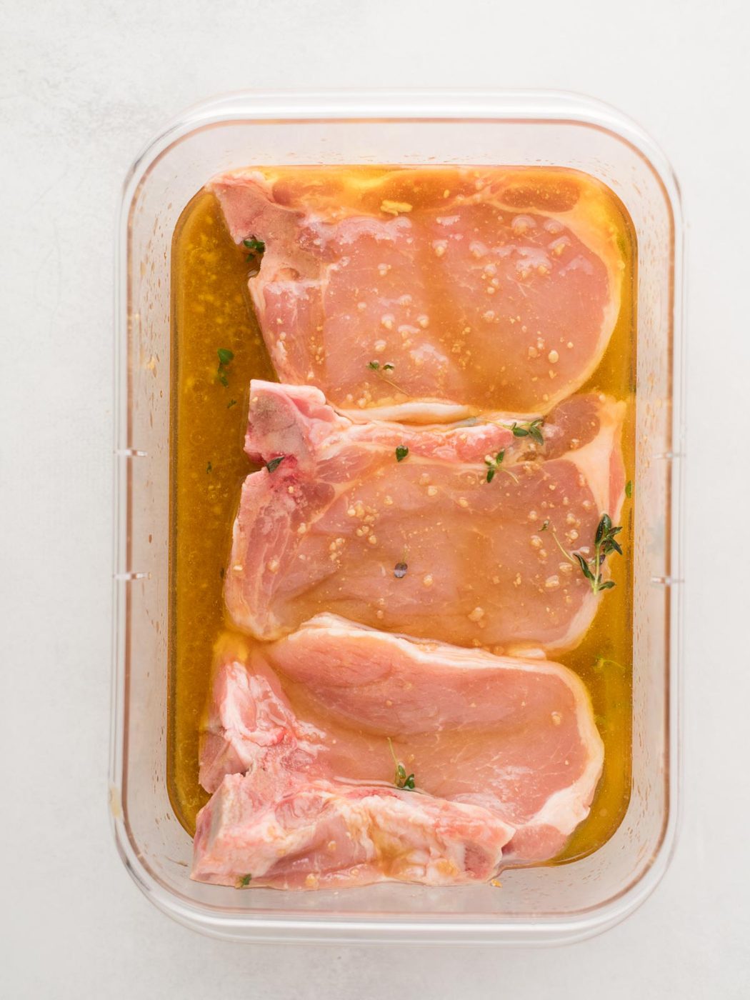 Three raw pork chops, soon to become flavorful grilled pork chops, are marinating in a rectangular container filled with a light brown marinade, garnished with small bits of herbs and garlic. The container sits on a white surface.