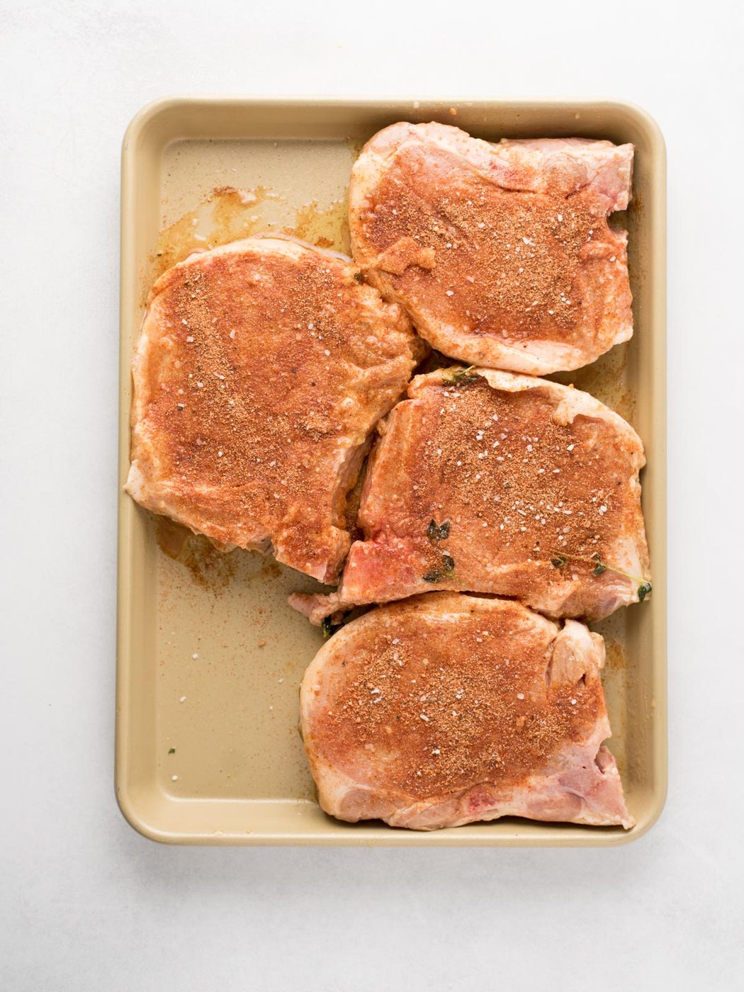 A baking tray with four raw pork chops sprinkled with seasoning, arranged on a light surface—evenly prepped for satisfying grilled pork chops.
