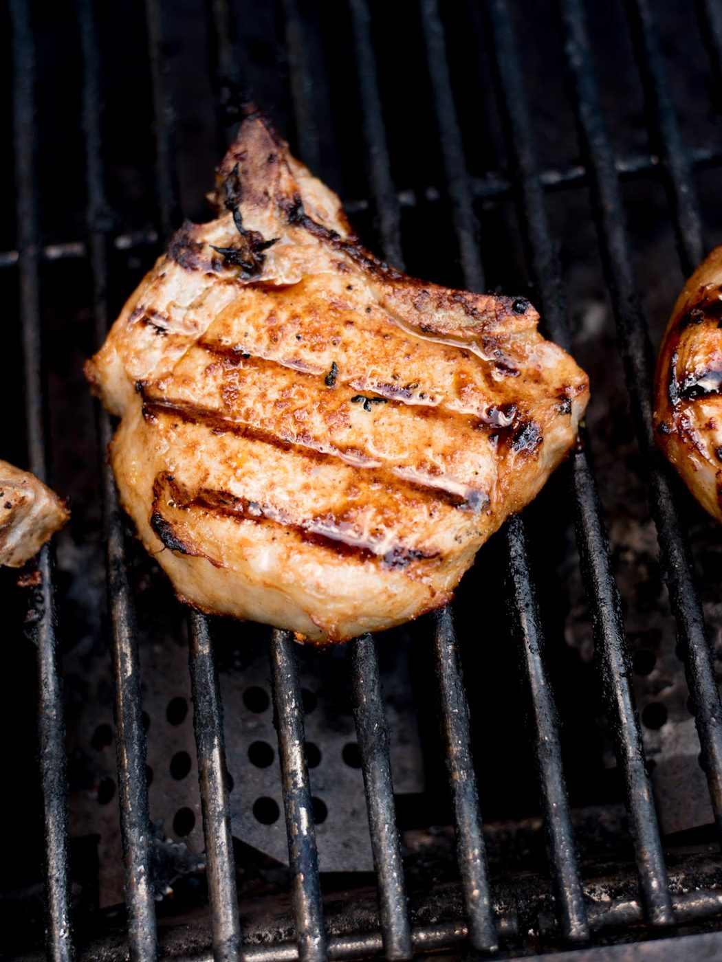 A grilled pork chop with visible grill marks cooks on a metal barbecue grill, showcasing charred lines and a golden-brown exterior—an irresistible example of evenly grilled pork chops.