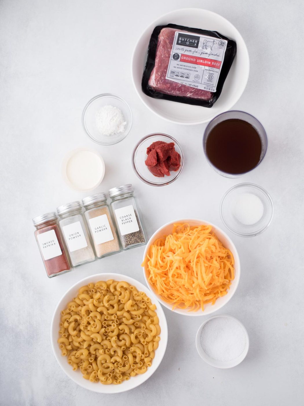 Bowls and jars with ground beef, shredded cheddar cheese, elbow macaroni, tomato paste, broth, cornstarch, sugar, and spices are neatly arranged on a white surface, viewed from above—everything you need to make a satisfying from-scratch hamburger helper.