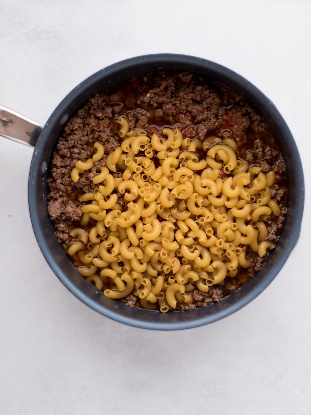 A pot filled with cooked ground beef and uncooked elbow macaroni pasta, ready to be mixed together for a from-scratch hamburger helper, sits on a white surface.