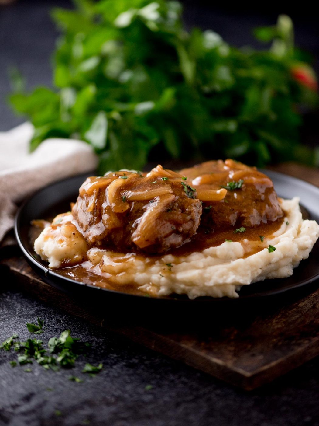 hamburger steak with gravy over a pile of mashed potatoes sitting on a black plate. The plate is on top of a wooden board with parsley in the background.