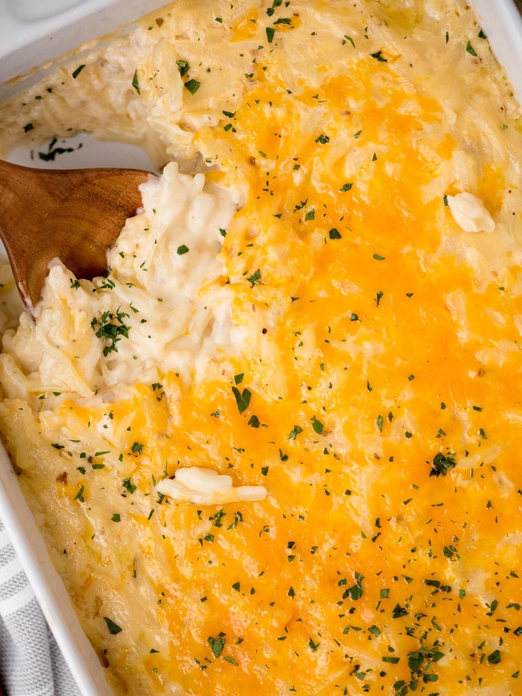 Close-up of a hash brown casserole in a white baking dish, with a wooden spoon lifting out a portion. The top layer is golden brown with melted cheese and garnished with parsley.