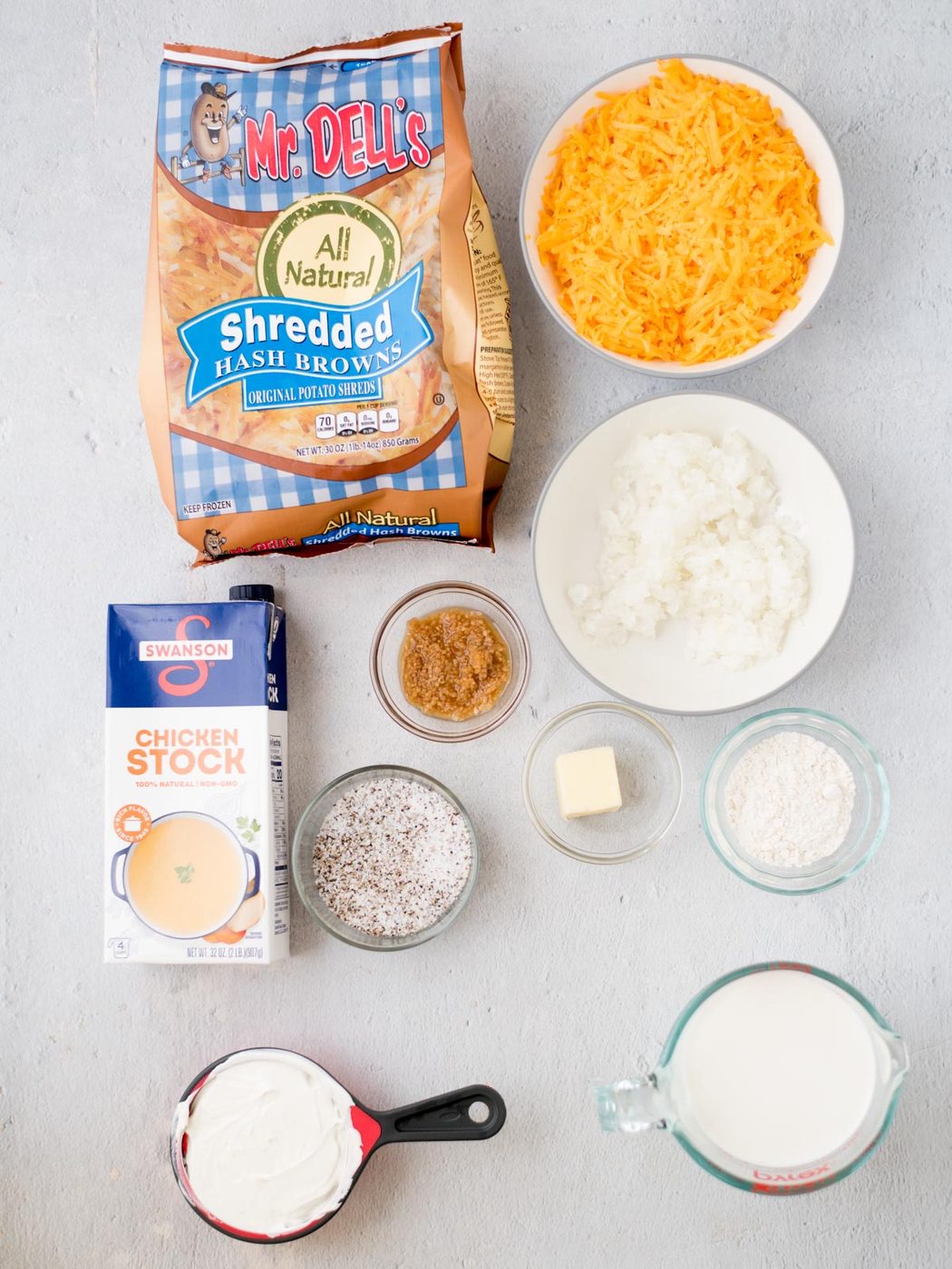 A collection of ingredients laid out for making hash brown casserole, including a bag of shredded hash browns, a bowl of shredded cheese, diced onions, a carton of chicken stock, a cup of sour cream, and small bowls containing various seasonings and butter.