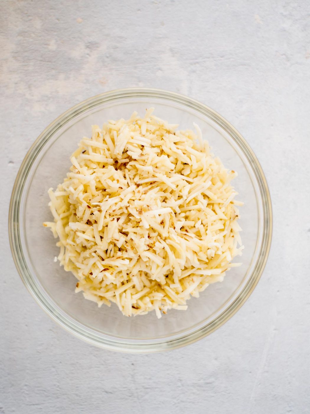 A glass mixing bowl filled with shredded hash browns, ready to be combined with other ingredients for making hash brown casserole.