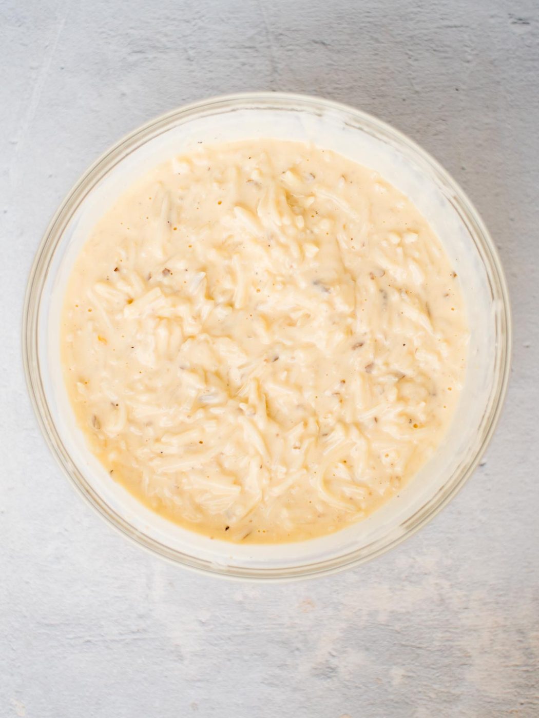 A glass mixing bowl containing hash browns mixed with a creamy cheese sauce, evenly coating the potatoes, ready to be transferred to a baking dish.