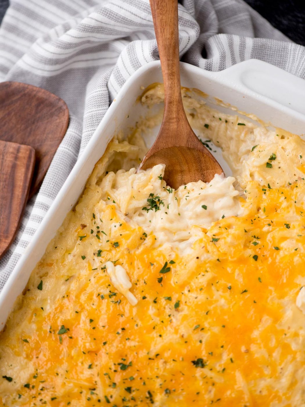 A wooden spoon scooping out a portion of creamy, cheesy hash brown casserole from a white baking dish. The top is golden and sprinkled with parsley.