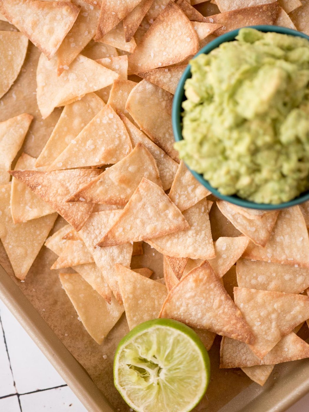 Small bowl of gucamole nestled in a tray of baked tortilla chips with a squeezed lime half.