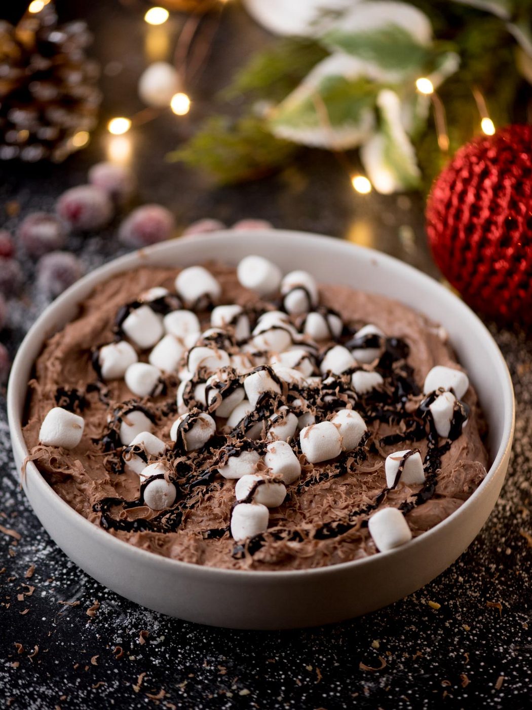 A bowl of hot cocoa dip topped with mini marshmallows and a drizzle of chocolate sauce. Festive decorations, including a red ornament and pinecones, are blurred in the background, along with twinkling lights.