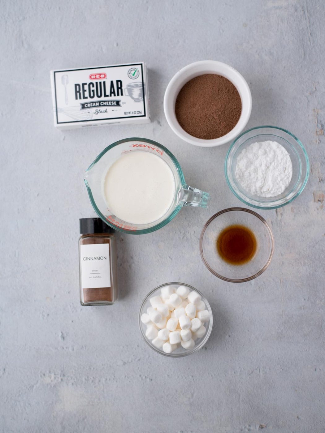 Ingredients on a gray surface include cream cheese, cocoa powder, powdered sugar, vanilla extract, cinnamon, and mini marshmallows.