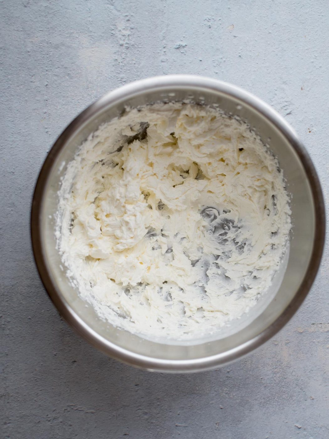 A stainless steel bowl filled with fluffy, whipped cream sits on a light gray textured surface. The cream appears smooth and airy, with visible whisk marks.