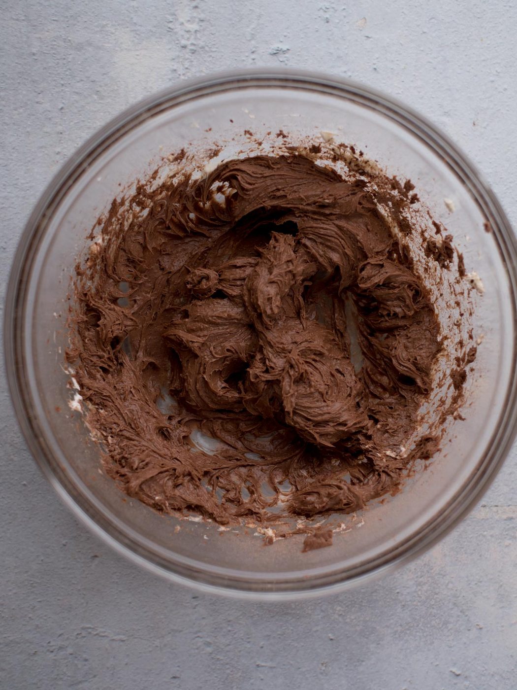 A clear glass bowl filled with rich, creamy chocolate dip on a light gray surface. The dip has a smooth, whipped texture with peaks and swirls visible on top.