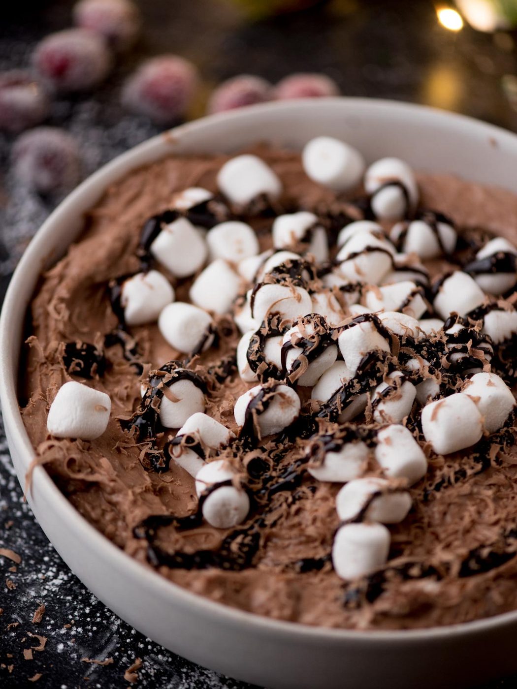 A bowl of chocolate dip topped with mini marshmallows and drizzled with chocolate sauce. The dessert is placed on a dark surface sprinkled with chocolate shavings, and there are blurred frozen berries in the background.