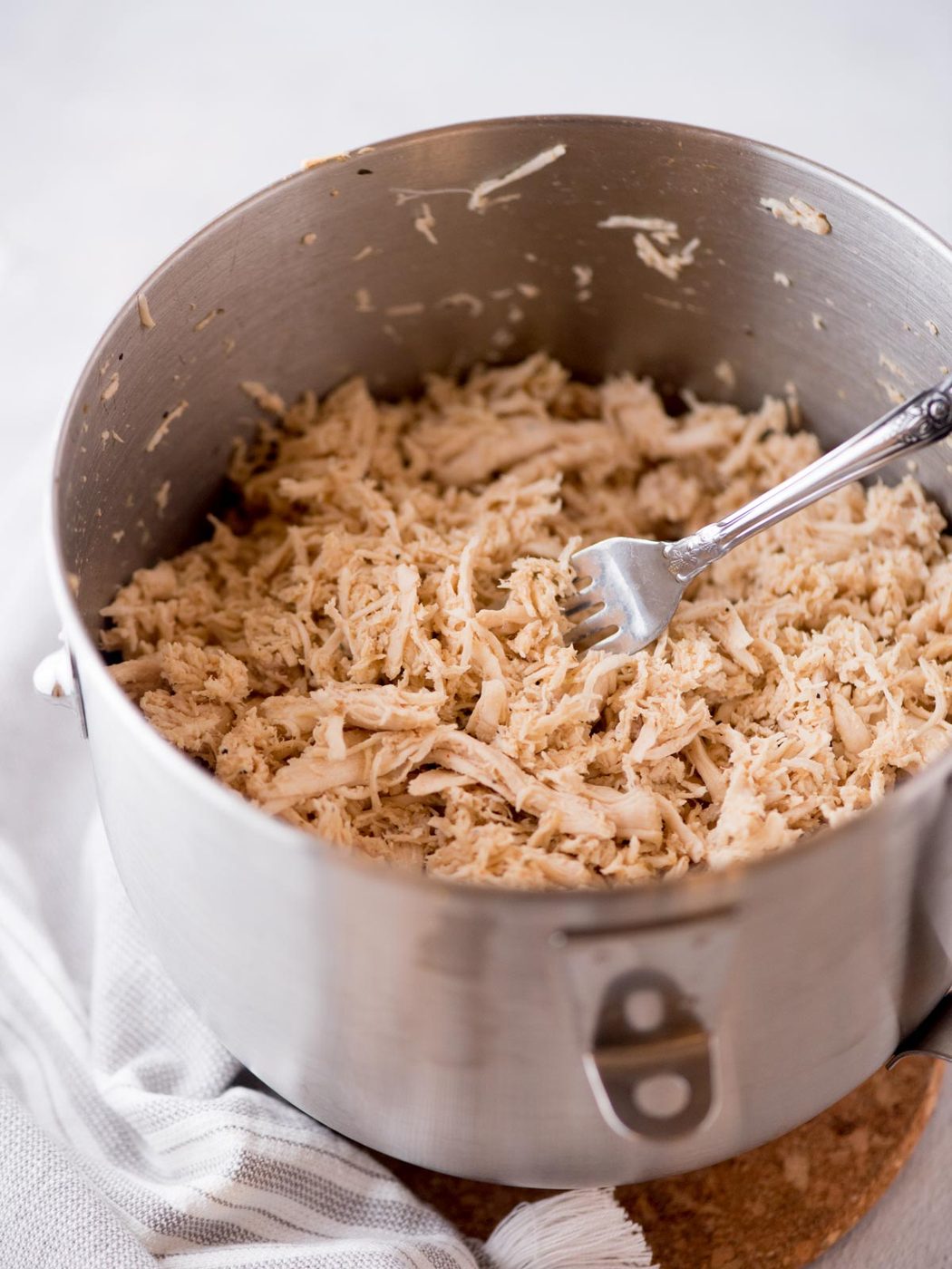 A large metal pot filled with shredded chicken in the Instant Pot, with a fork resting inside. The pot sits on a cork trivet atop a light-colored cloth.